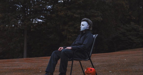 A figure in a white mask sits on a chair in a dark, wooded area, with a carved pumpkin nearby.