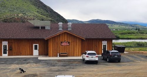 A wooden building with a sign, surrounded by mountains and a river, with cars parked outside and a dog on the ground.