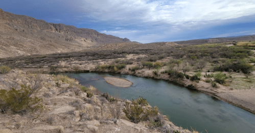 A winding river flows through a desert landscape with mountains and sparse vegetation under a cloudy sky.