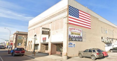 A building with a large American flag mural, featuring signs for a fertilizer supply and a "For Sale" notice.