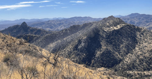 A panoramic view of rugged mountains and valleys under a clear blue sky, with dry grass in the foreground.