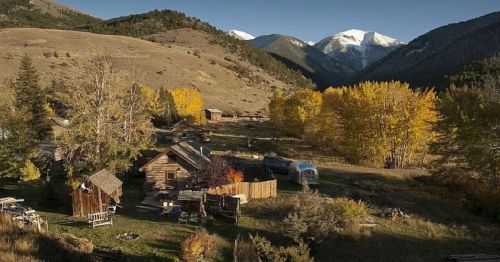 A scenic view of a rustic settlement surrounded by mountains and autumn trees, with snow-capped peaks in the background.
