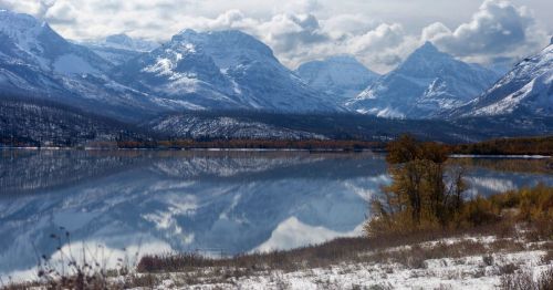 A serene lake reflects snow-capped mountains and cloudy skies, surrounded by autumn foliage and a snowy shoreline.