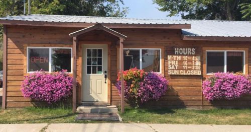 A wooden building with a welcoming entrance, surrounded by vibrant pink flowers and a sign displaying business hours.