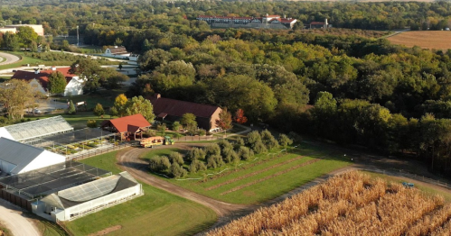 Aerial view of a farm with greenhouses, fields, and a red-roofed building surrounded by trees and farmland.