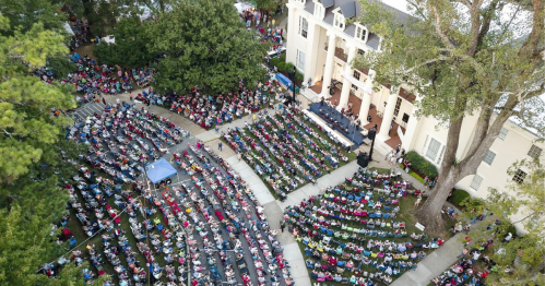Aerial view of a large outdoor crowd seated in rows, gathered for an event in front of a historic building.