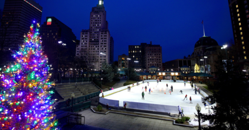 A vibrant holiday tree stands beside an ice rink, with skaters enjoying the evening in a cityscape backdrop.