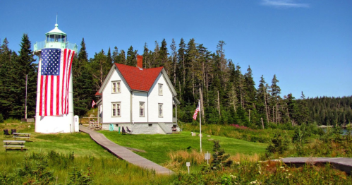A lighthouse with an American flag, next to a white house, surrounded by green grass and trees under a clear blue sky.