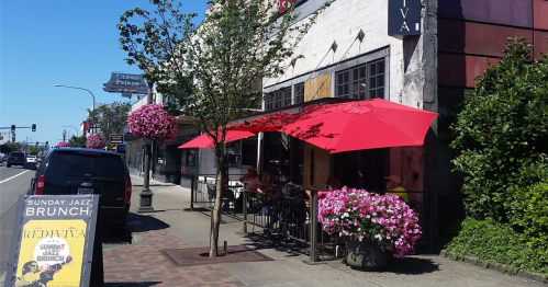 A street view of a restaurant with red umbrellas, flowers, and people dining outside on a sunny day.