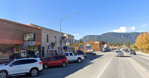 A street view of a small town with a café, shops, and mountains in the background under a clear blue sky.