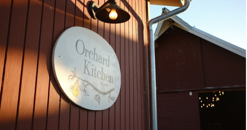 Sign for "Orchard Kitchen" on a red barn wall, with a light fixture above it.