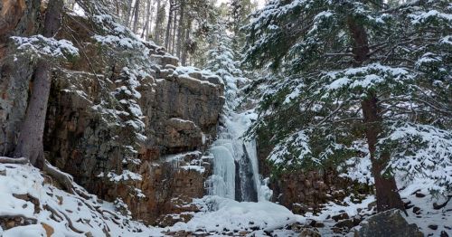 A snowy waterfall cascading down rocky cliffs, surrounded by tall evergreen trees in a winter landscape.