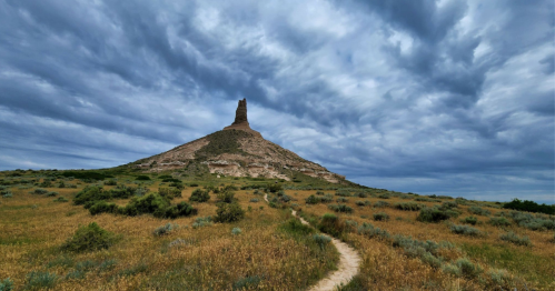 A rocky butte rises from a grassy landscape under a dramatic, cloudy sky. A winding path leads toward the formation.