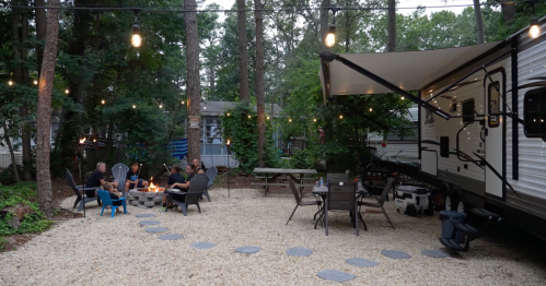 A cozy outdoor space with a fire pit, seating area, and an RV surrounded by trees and string lights.