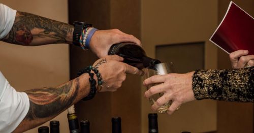 A person pours wine from a bottle into a glass held by another person, with wine bottles visible in the background.