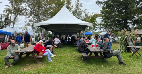 A gathering at an outdoor event with people sitting at picnic tables under a large tent, surrounded by trees.
