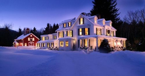 A snowy landscape featuring a beautifully lit white house and a red barn, surrounded by trees at dusk.