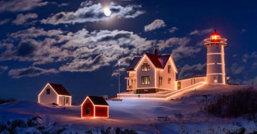 A snowy landscape at night featuring a lighthouse and house adorned with festive lights under a full moon.