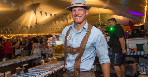 A man in a hat and suspenders holds a large beer mug at a festive outdoor event with decorations and people in the background.