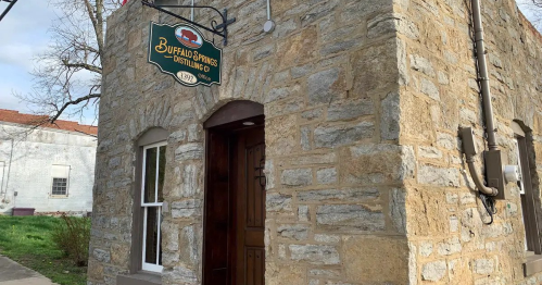 Stone building with a wooden door and a sign reading "Buffalo Springs Distilling Co." in a quaint setting.