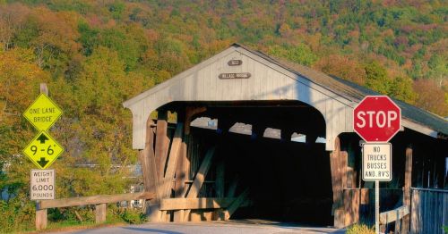 A rustic covered bridge with a stop sign and weight limit signs, surrounded by colorful autumn foliage.