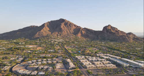 Aerial view of a mountainous landscape with residential areas and greenery in the foreground. Clear blue sky above.