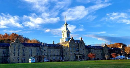 Historic building with a tall spire, surrounded by trees and a blue sky with clouds. Green lawn in the foreground.