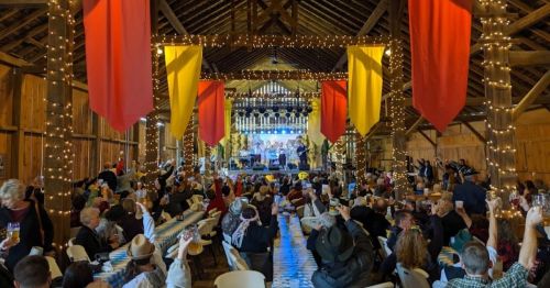 A lively barn event with people raising glasses, colorful banners, and a band performing on stage, decorated with string lights.