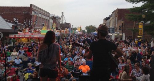A large crowd enjoys a street festival, with food stalls and a live performance in the foreground.