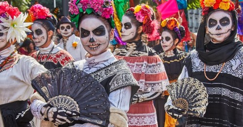 A group of people in traditional attire with skull makeup and colorful flowers, holding fans during a festive celebration.