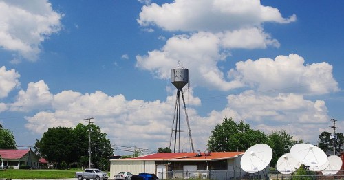 A water tower stands tall against a blue sky with fluffy clouds, near buildings and satellite dishes.