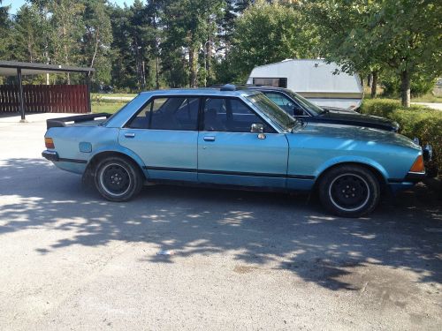 A vintage blue sedan parked in a lot, surrounded by trees and another car in the background.