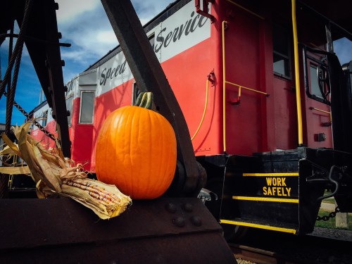 A bright orange pumpkin and dried corn sit on a train car, with a "Work Safely" sign visible in the background.