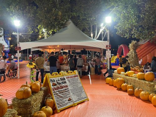 A festive outdoor scene with a tent, pumpkins, and people enjoying a fall event under bright lights.