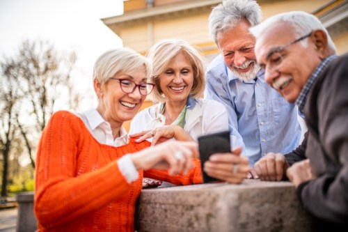 Four smiling adults gather around a smartphone, enjoying a moment together outdoors.