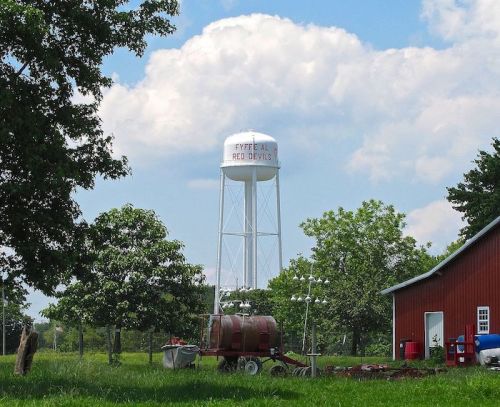 Water tower labeled "Pyrite All Red Devil" beside a red barn and green trees under a blue sky with clouds.