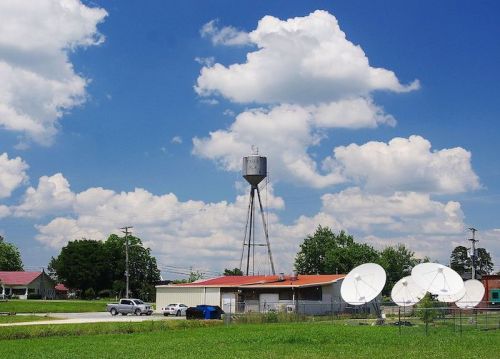 A water tower stands tall against a blue sky with clouds, near a building and satellite dishes in a grassy area.