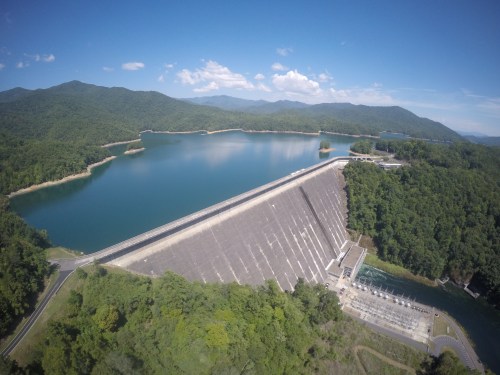 Aerial view of a large dam surrounded by lush green mountains and a calm blue lake under a clear sky.