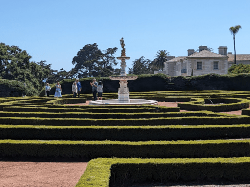 A garden maze with a fountain at the center, surrounded by visitors and a historic building in the background.