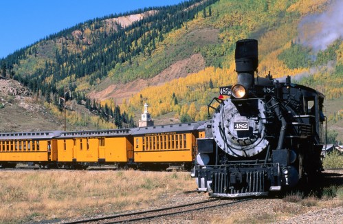 A vintage steam train with yellow carriages travels through a scenic landscape of mountains and autumn foliage.
