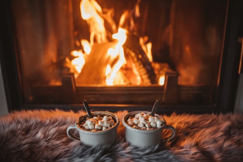 Two mugs of hot chocolate topped with marshmallows sit in front of a cozy fireplace, with a soft fur blanket underneath.