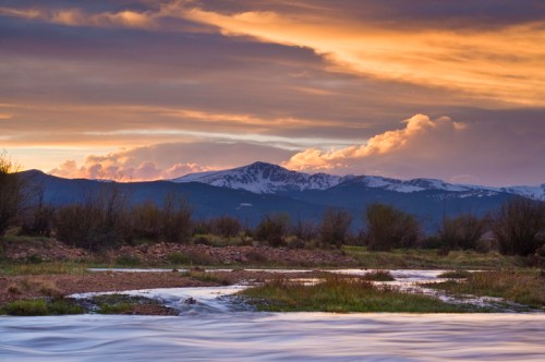 A serene landscape featuring a river, grassy banks, and mountains under a colorful sunset sky with clouds.