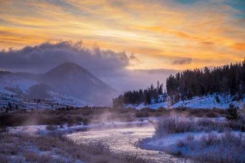 A serene winter landscape with a river, mountains, and colorful sunset skies, surrounded by frosty trees and mist.
