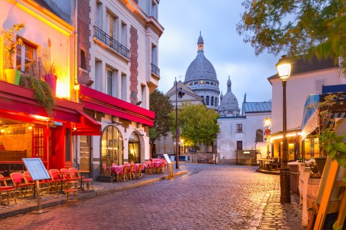 Charming Parisian street at dusk, featuring cafes, cobblestones, and the Basilica of Sacré-Cœur in the background.
