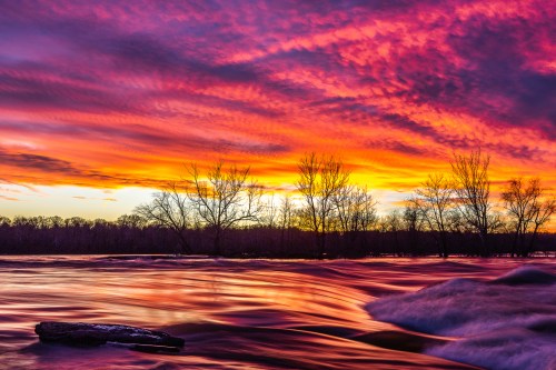 A vibrant sunset over a river, with colorful clouds and silhouetted trees along the shoreline.