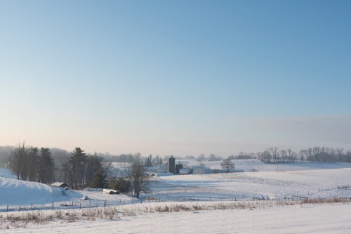 A serene winter landscape featuring snow-covered fields, trees, and a farm with silos under a clear blue sky.