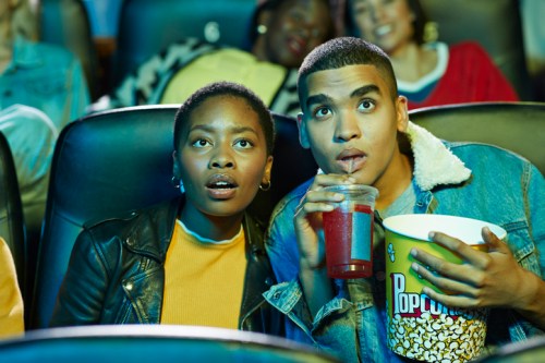 A young couple watches a movie in a theater, looking surprised while holding popcorn and a drink.