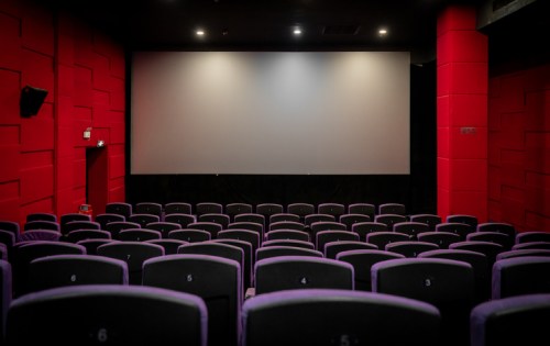 Empty cinema hall with purple seats and a large blank screen, illuminated by soft overhead lights.