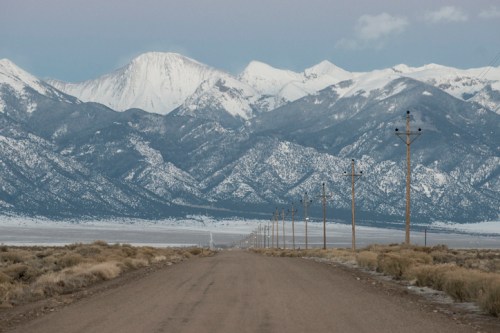 A dirt road stretches towards snow-capped mountains, with power lines running alongside in a serene landscape.