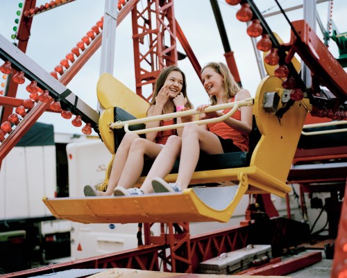 Two girls sit in a yellow Ferris wheel seat, smiling and enjoying ice cream at a fairground.